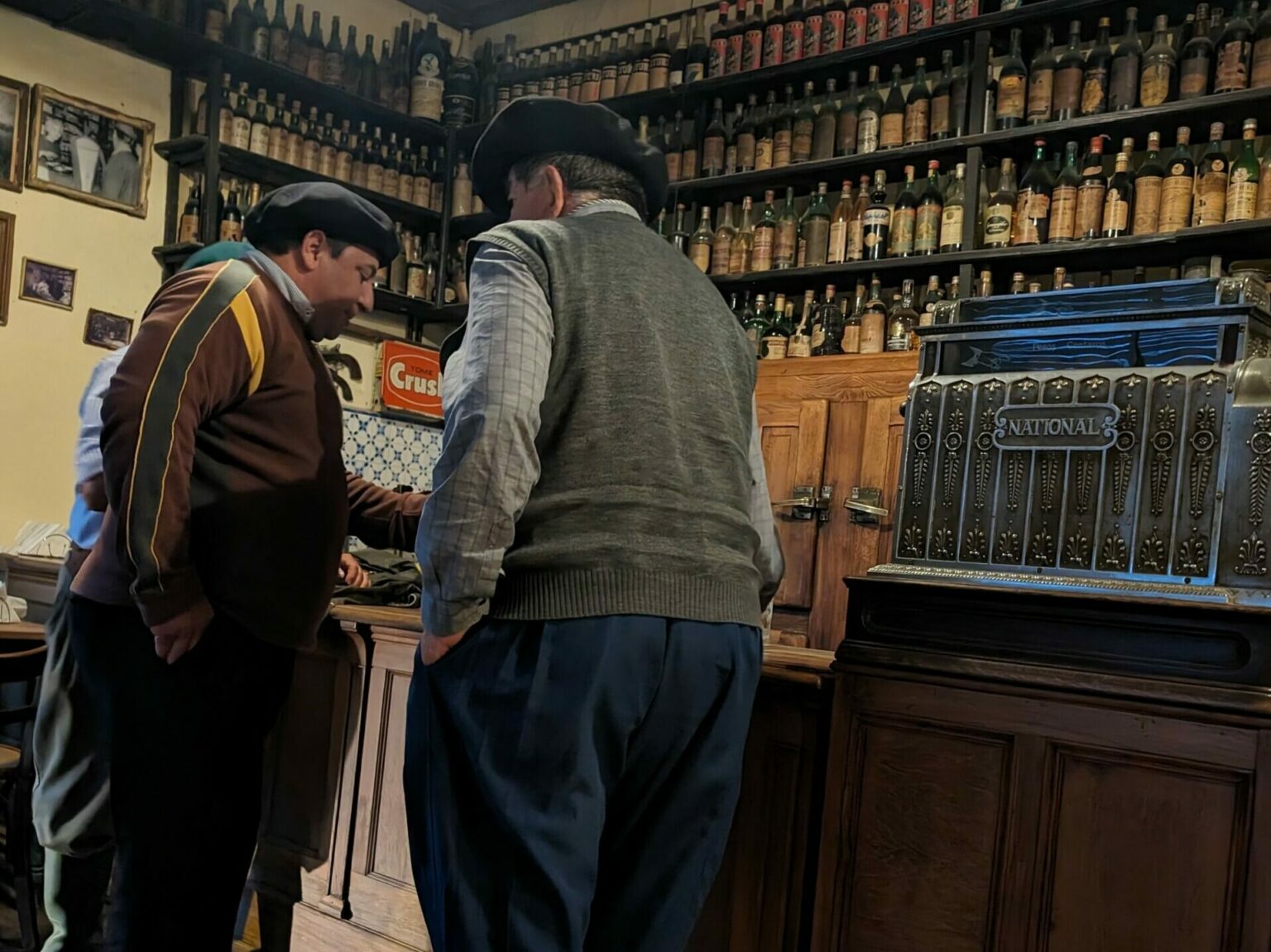 Two men in traditional dress standing at a bar with bottles in the background.