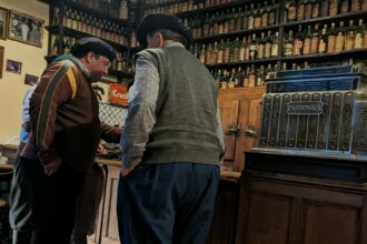 Two men in traditional dress standing at a bar with bottles in the background.