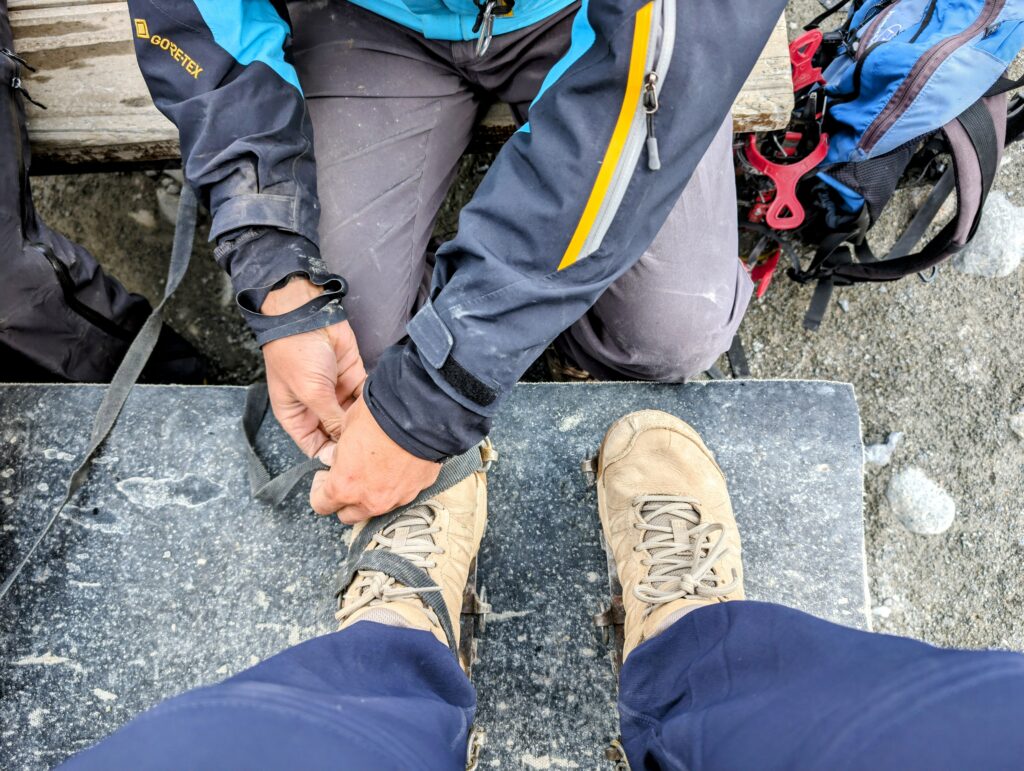 A person attaching crampons on the shoes of another person, seen from above.