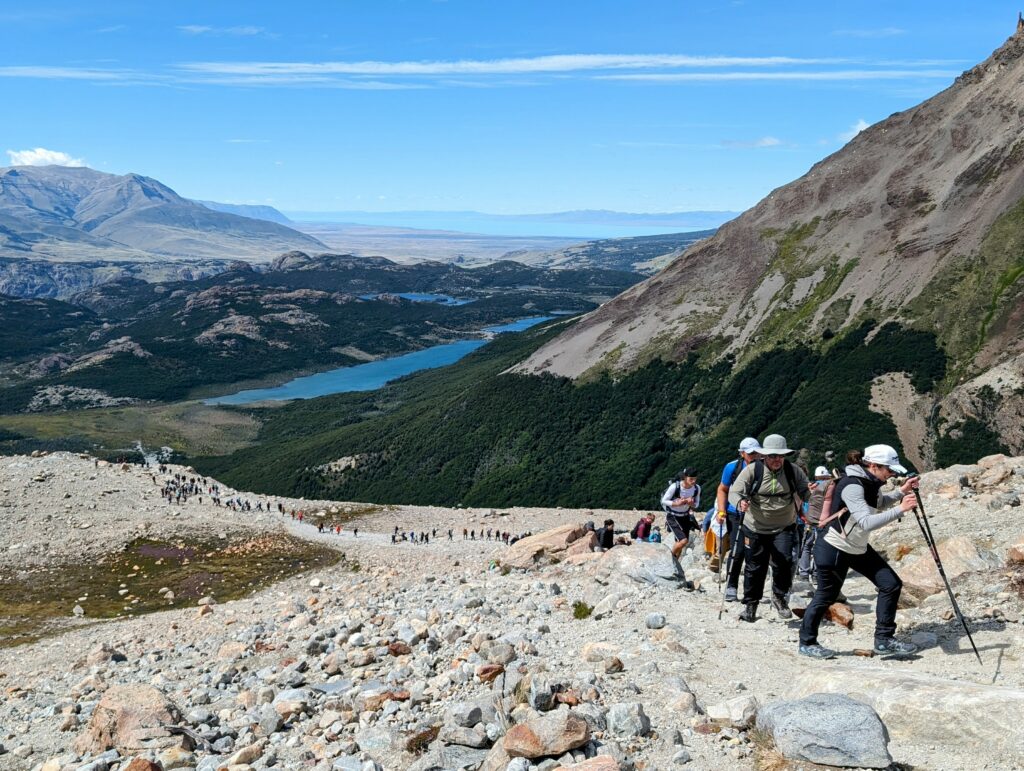A line of people hiking up a mountain with a lake and valley in the distance