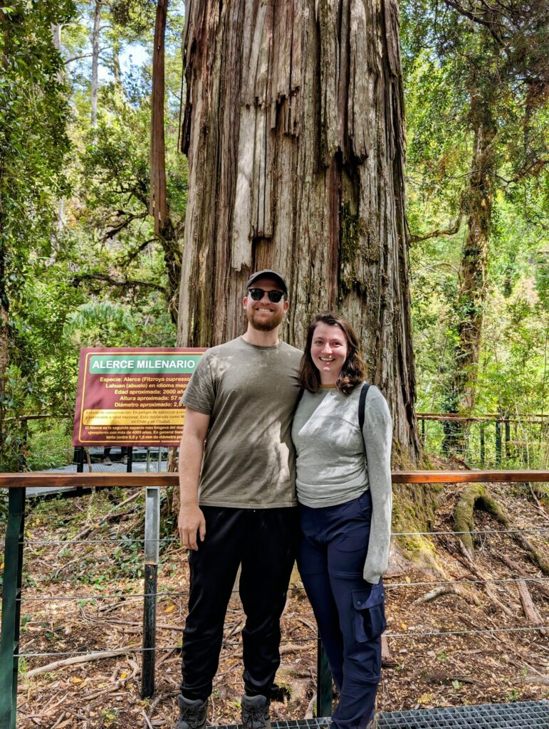 Two people smiling standing in front of a large tree in a forest