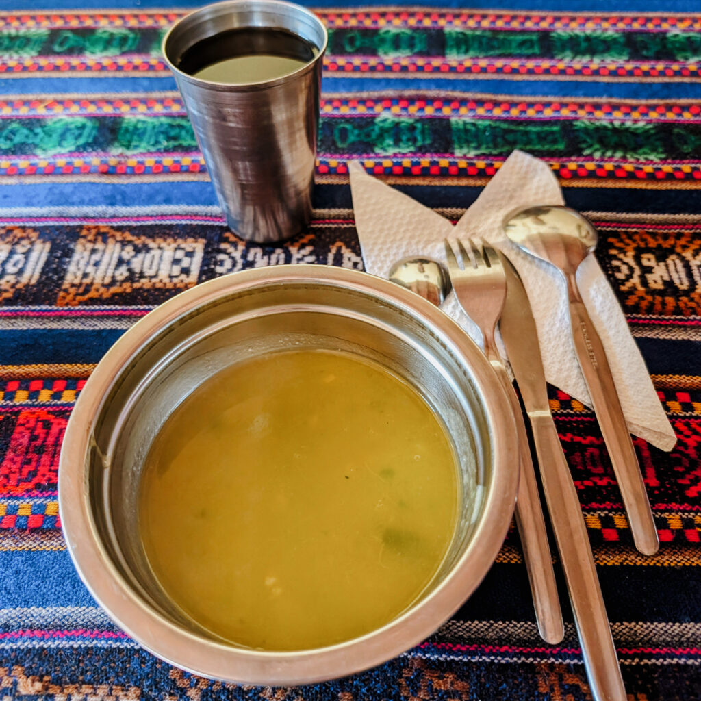 A vegetable soup in a metal bowl with cutlary on the side and a cup of juice for a meal on the Lares Trek