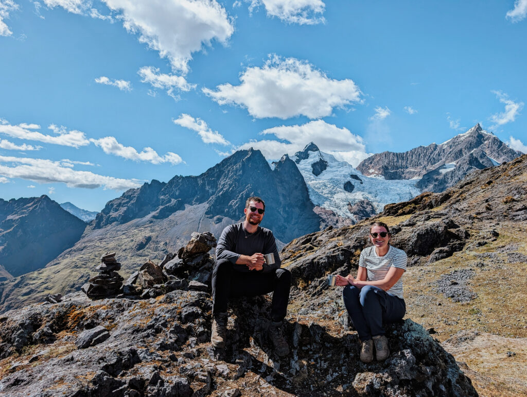Two people sitting on a rock with mugs in their hands in front of a snow-covered mountain on the Lares Trek