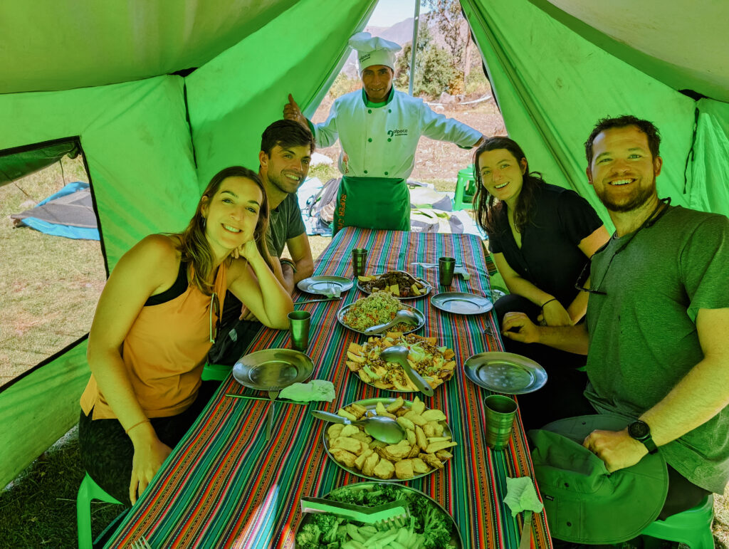 People sitting around a table full of food with a chef in the back under a green tent during the Lares Trek