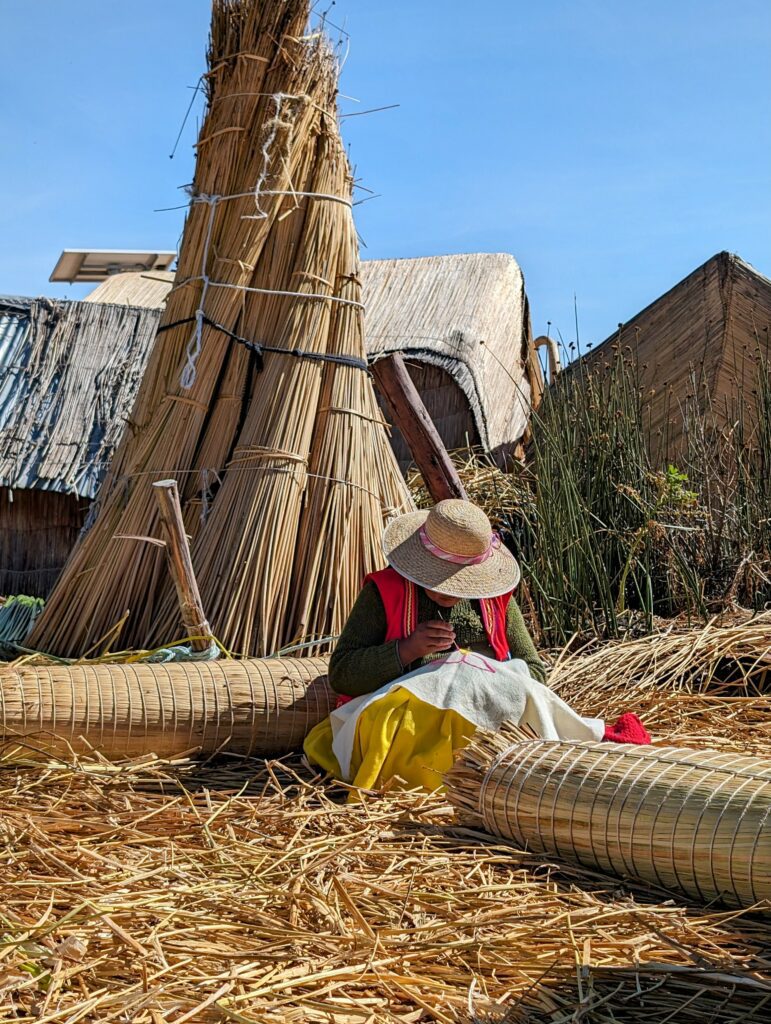 A woman embroidering in front of a pile of reeds on land covered in reeds on a Lake Titicaca homestay tour