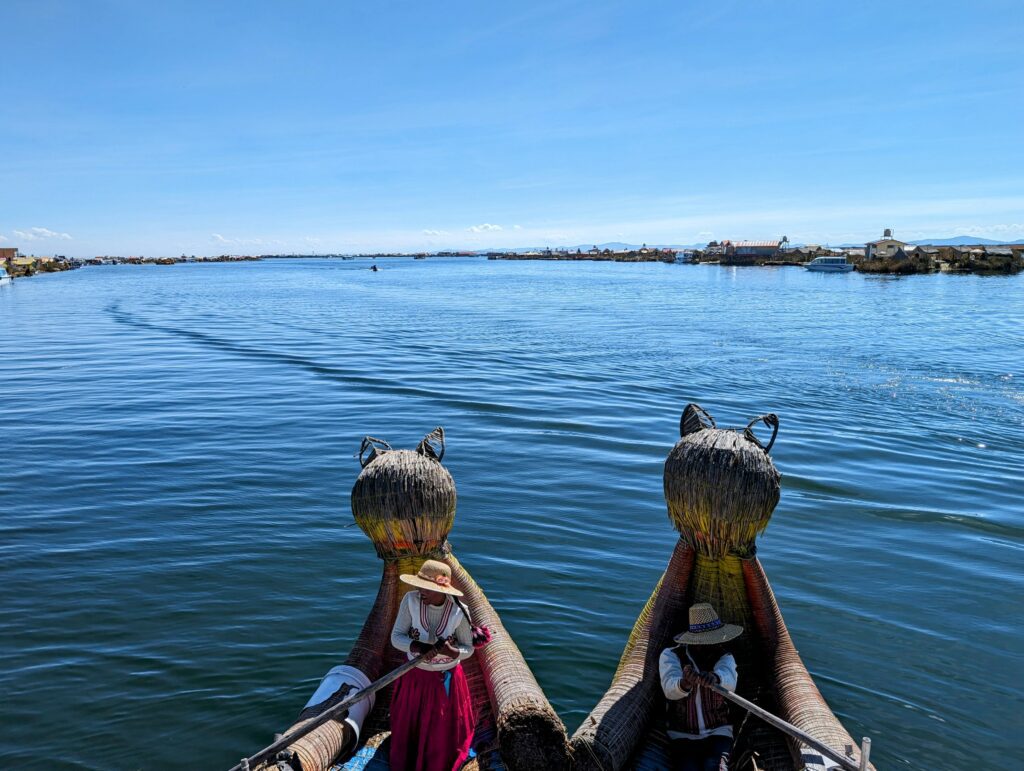 Two people rowing a boat made of reeds with cat heads at the top of the boat on a Lake Titicaca homestay tour