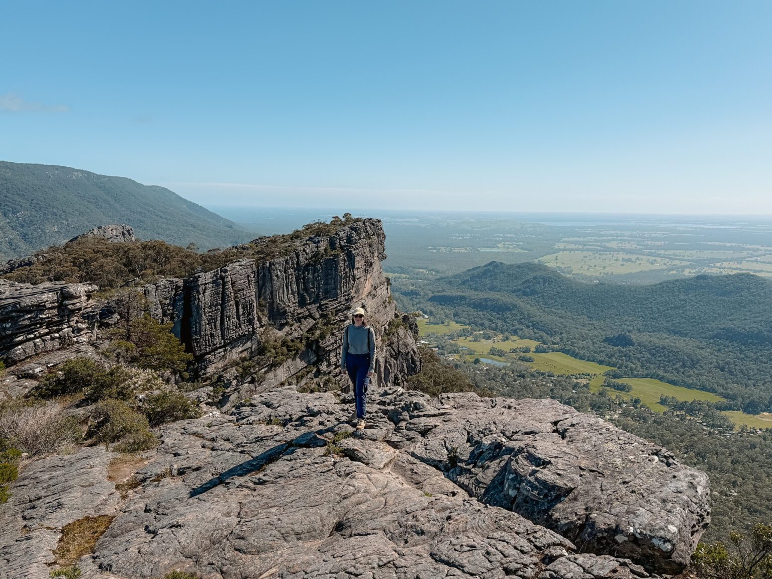 A woman standing in front of a rock at the peak of a small mountain overlooking a green valley in Grampians