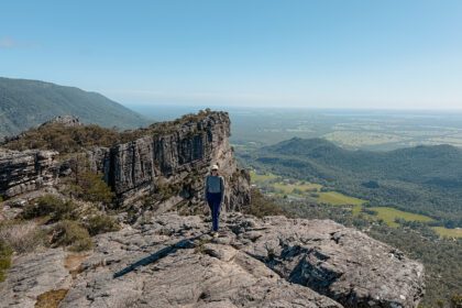 A woman standing in front of a rock at the peak of a small mountain overlooking a green valley in Grampians